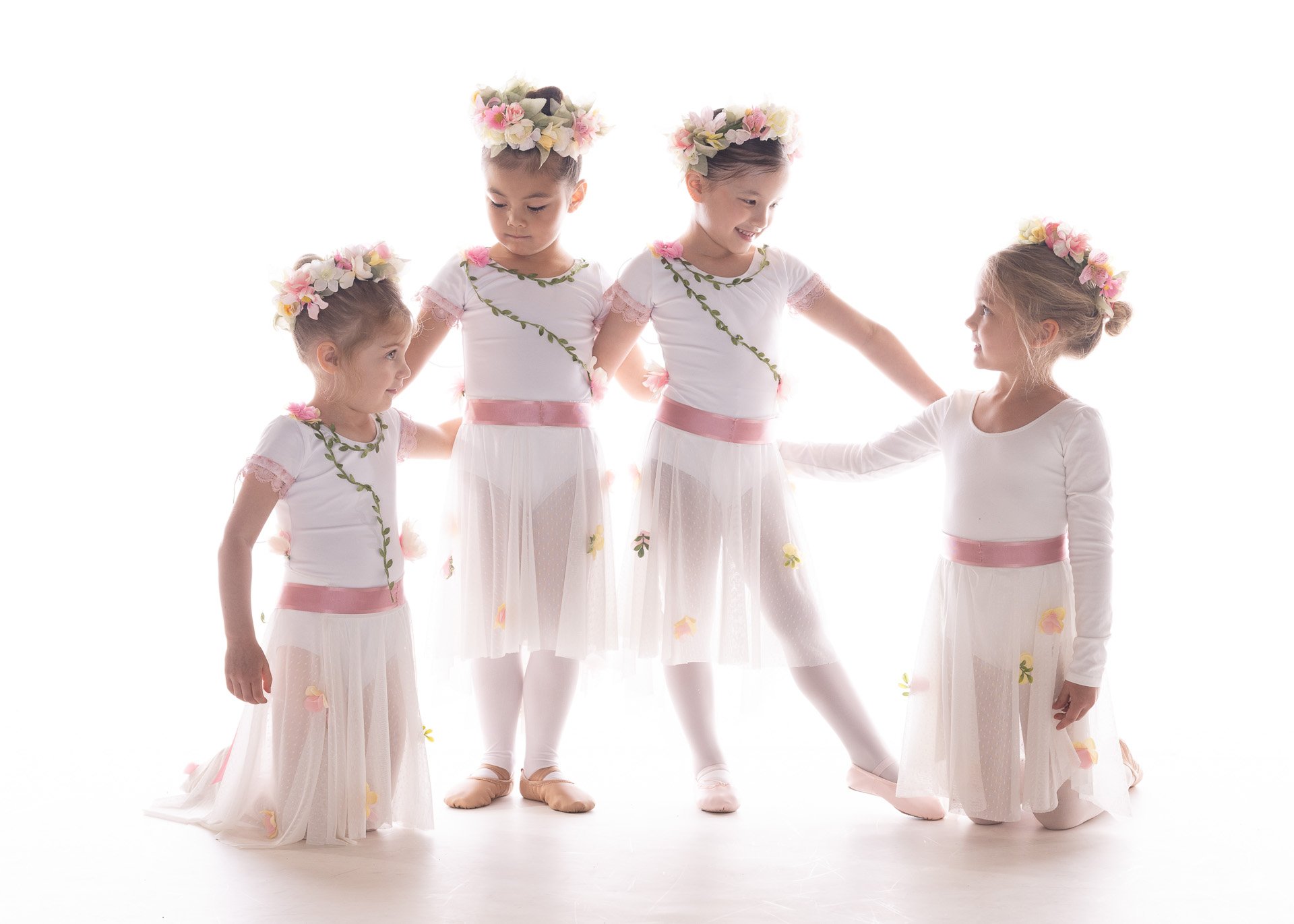 Four young ballet dancers in white dresses holding hands in a bright studio